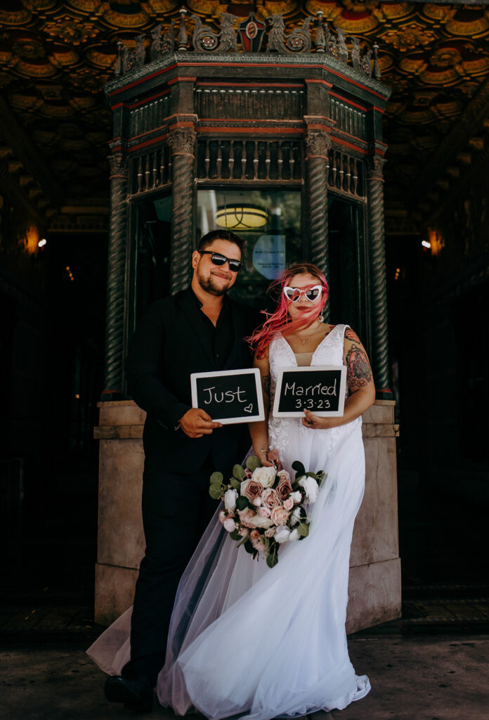 bride and groom after their tampa courthouse wedding and are standing in their wedding attire with a sign that says just married in front of the ticket booth of a theatre 